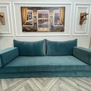 May include: A teal blue futon sofa with two large pillows and two armrests. The sofa is folded out and ready for use. The sofa is in front of a white wall with a framed picture of a city skyline above it.