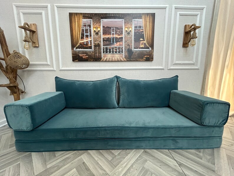 May include: A teal blue futon sofa with two large pillows and two armrests. The sofa is folded out and ready for use. The sofa is in front of a white wall with a framed picture of a city skyline above it.