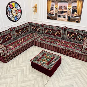 May include: A red and gold patterned sectional sofa with a matching red square ottoman. The sofa is made up of four individual pieces, each with a cushion and a patterned fabric. The sofa is set against a white wall with a clock and a window in the background.
