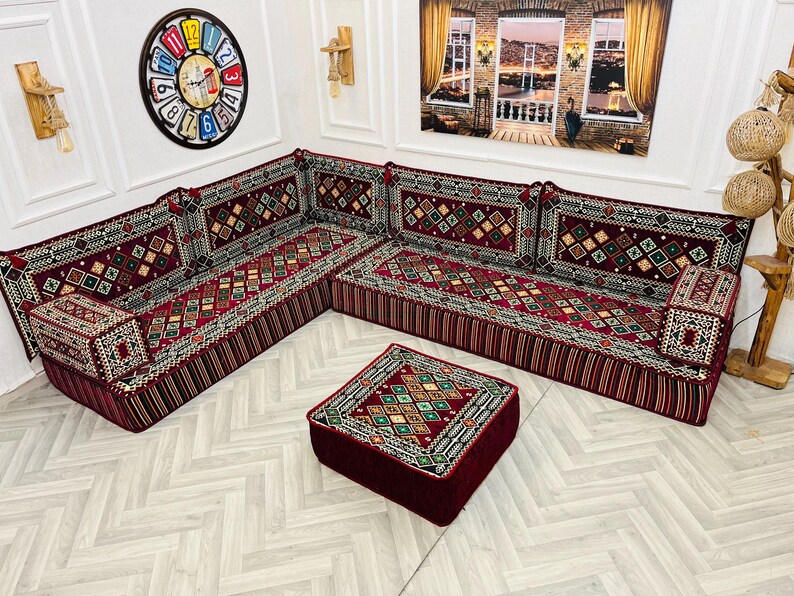 May include: A red and gold patterned sectional sofa with a matching red square ottoman. The sofa is made up of four individual pieces, each with a cushion and a patterned fabric. The sofa is set against a white wall with a clock and a window in the background.