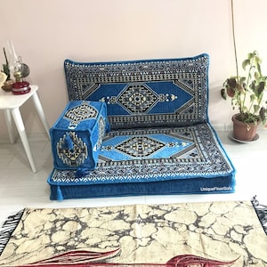May include: A blue floor sofa set with a patterned design. The set includes a backrest, a seat cushion, and an armrest. The design features geometric shapes in shades of blue, black, and white. Tassels adorn the armrest. A patterned rug is in the foreground.