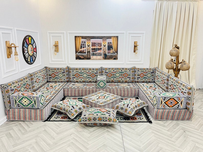 May include: A gray and white sectional sofa with colorful patterned cushions. The sofa is arranged in a U-shape and has a variety of throw pillows in different patterns and colors. The sofa is set against a white wall with a window behind it.