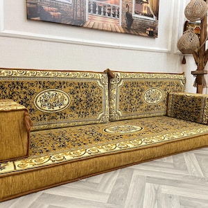 May include: A traditional Middle Eastern floor seating set with a patterned fabric in shades of brown and gold. The set includes a long, rectangular cushion with two backrests and a smaller square cushion.