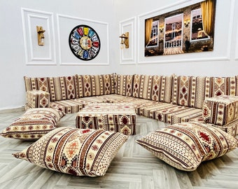 May include: A traditional Middle Eastern seating arrangement with a corner sofa and several cushions. The sofa and cushions are covered in a beige fabric with a brown and red geometric pattern. There is a small coffee table in the center of the seating area.