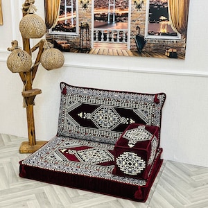 Puede incluir: Un juego de cojines de suelo burdeos con un estampado geométrico en blanco, negro y dorado. El juego incluye un cojín de suelo grande, un respaldo y dos cojines cuadrados más pequeños. Los cojines tienen borlas en las esquinas.