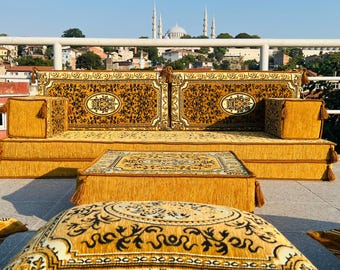 May include: A gold and brown patterned sofa with matching cushions and a coffee table. The sofa is made of fabric and has a traditional design. The sofa is set against a backdrop of a city skyline with a mosque in the distance.