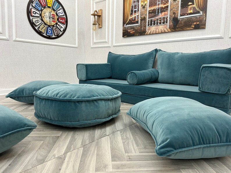 May include: A teal blue sofa with matching throw pillows and floor cushions. The sofa has a low back and is perfect for lounging. The floor cushions are round and have a tufted design.
