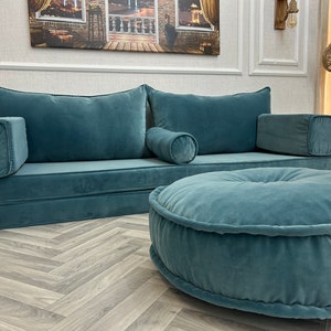 May include: A teal blue velvet sofa with a matching ottoman. The sofa has two large pillows and a smaller pillow in the center. The ottoman is round and has a tufted design.