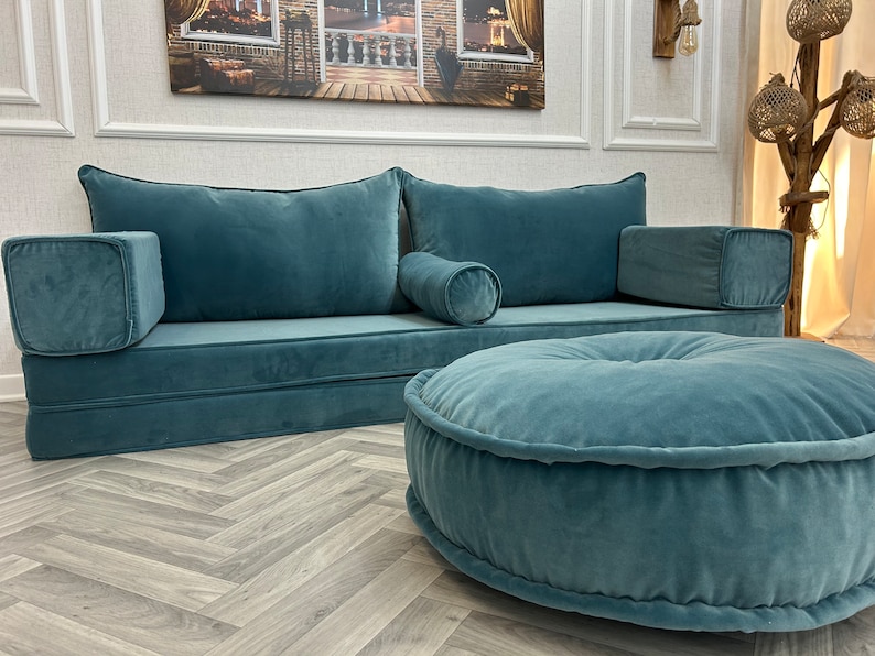 May include: A teal blue velvet sofa with a matching ottoman. The sofa has two large pillows and a smaller pillow in the center. The ottoman is round and has a tufted design.