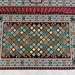 May include: A red, black, and green patterned rug with a geometric design. The rug has a fringe border and is made of a soft, plush material.
