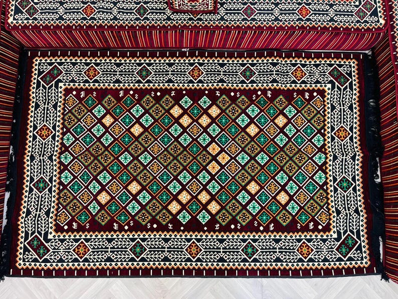 May include: A red, black, and green patterned rug with a geometric design. The rug has a fringe border and is made of a soft, plush material.