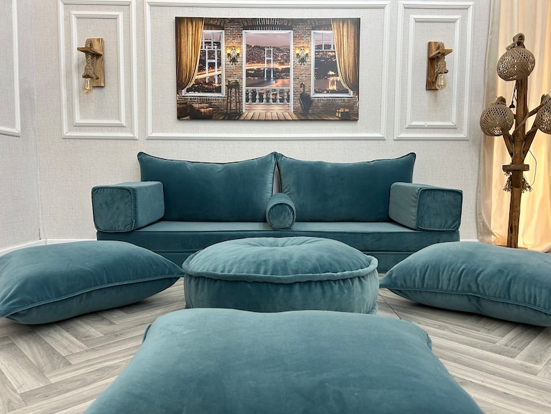 May include: A teal blue sofa with matching floor cushions and pillows. The sofa has a low back and armrests. The cushions are arranged in a symmetrical pattern on the floor in front of the sofa.