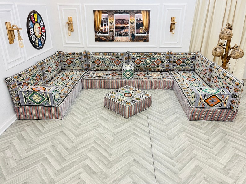 May include: A traditional gray and white patterned divan with a matching ottoman. The divan is made up of multiple sections and has a variety of colorful geometric patterns. The divan is set on a light gray wood floor.