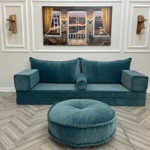May include: A teal blue sofa with two large pillows and a smaller pillow in the center. The sofa is in a room with a wood floor and a painting on the wall. There is a teal blue round ottoman in front of the sofa.