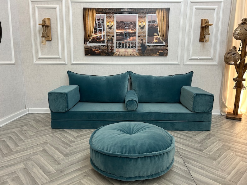 May include: A teal blue sofa with two large pillows and a smaller pillow in the center. The sofa is in a room with a wood floor and a painting on the wall. There is a teal blue round ottoman in front of the sofa.