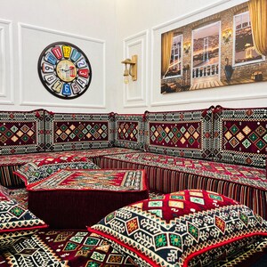 May include: A red and gold patterned floor cushion set with a variety of cushions and pillows. The cushions are arranged in a traditional seating style, with a large central cushion and smaller cushions around it. The cushions are made of a soft, plush fabric and have a variety of intricate patterns.