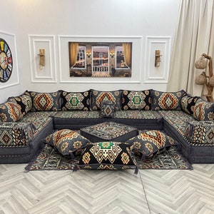 May include: A large, grey, L-shaped sectional sofa with multiple cushions. The cushions are covered in a black and brown geometric patterned fabric. The sofa is set on a patterned rug with a small, square, grey ottoman in the centre.