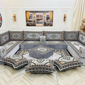 May include: A grey and white patterned floor seating set with a grey and white patterned rug. The set includes a large couch with multiple cushions, a small square ottoman, and several throw pillows. The couch is arranged in a U-shape.