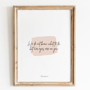 Op de afbeelding: Een ingelijst print met een lichtroze achtergrond en de tekst "We do not know what to do, but our eyes are on you." De tekst is in zwart cursief schrift. De print is ingelijst in een lichtbruin houten lijst.