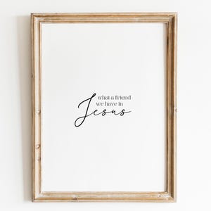Op de afbeelding: Een ingelijst afdruk met een witte achtergrond en zwarte tekst die "Wat een vriend hebben we in Jezus" luidt.