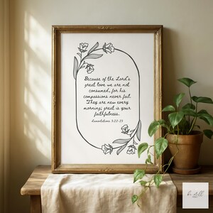 May include: A framed print with a gold-coloured frame. The print features a black and white floral design surrounding text from Lamentations 3:22-23. The text reads, "Because of the Lord's great love we are not consumed..." A potted plant sits nearby.