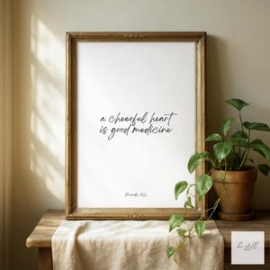 Może przedstawiać: Oprawiony wydruk z cytatem "a cheerful heart is good medicine" w czarnym skrypcie. Złota rama otacza biały nadruk, wystawiony na drewnianym stole z beżową szmatką. Po prawej stronie znajduje się roślina doniczkowa.