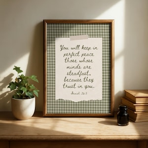 May include: A framed print with a quote from Isaiah 26:3, set against a green plaid background. The quote reads, "You will keep in perfect peace those whose minds are steadfast, because they trust in you." A small potted plant and books are on the wooden surface.