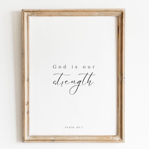 Puede incluir: Impresión enmarcada con las palabras "God is our strength" en una fuente elegante. La impresión está en un marco de madera clara e incluye el texto "Psalm 46:1" en la parte inferior. El fondo es blanco.