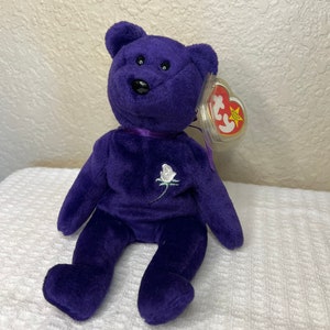 May include: Purple Beanie Baby bear with a white embroidered rose on its chest. The bear has black button eyes and a red Ty heart tag.