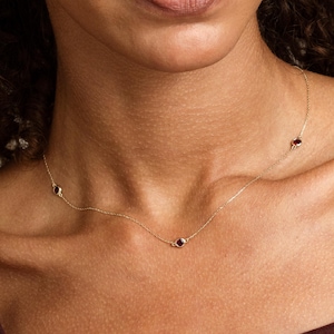 May include: A delicate gold necklace featuring three small, round, red gemstones. The necklace is displayed against a warm skin tone, highlighting the jewelry's subtle elegance. The necklace is a simple, yet sophisticated accessory.
