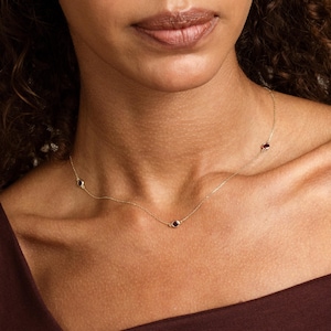 May include: A delicate gold chain necklace featuring evenly spaced, small, round, bezel-set dark red gemstones. The necklace rests against the neck, showcasing a minimalist design with a thin metallic link chain and multiple stone accents.