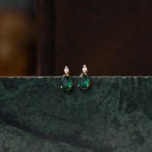 May include: A pair of gold stud earrings featuring a teardrop-shaped green gemstone topped with a small, round, clear white stone. The earrings are set in a gold-toned metal frame, showcasing a classic design with a vibrant green hue.