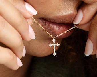 May include: A gold chain necklace featuring a cross pendant encrusted with small, clear, sparkling stones. The cross has a distinct shape with rounded, decorative ends on each of the four arms, suspended from a delicate, thin gold chain.