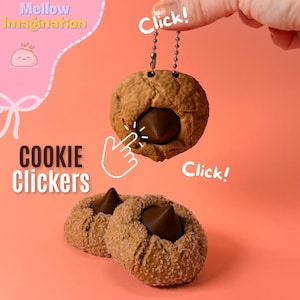 May include: A close-up of three cookie clickers on a peach background. The cookies are brown with a dark chocolate center. One cookie is attached to a chain. The text "Cookie Clickers" is in the lower left corner.