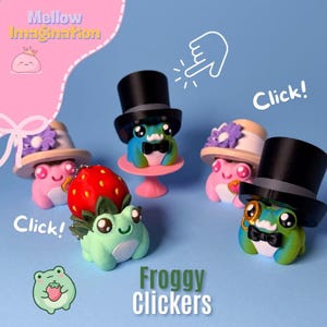 May include: A collection of colorful frog figurines, each with unique hats and accessories. The frogs are in shades of pink, blue, and green, with top hats, bow ties, and floral accents. The text "Froggy Clickers" is displayed.