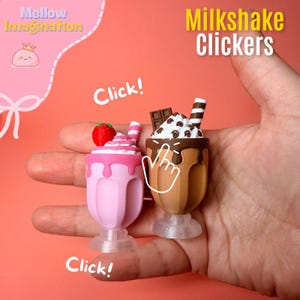 May include: Two miniature milkshake clickers, one pink with a strawberry and the other brown with chocolate toppings, held in a hand. The text "Milkshake Clickers" and "Click!" are visible. The background is a solid coral color.
