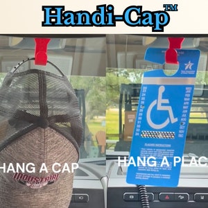 May include: A red plastic hook with a black cord hangs from the rearview mirror of a car. The hook is holding a brown and black baseball cap with a white logo that says "INDUSTRIAL" and a red star. The hook is also holding a blue placard with a white wheelchair symbol and the text "HANDI-CAP" in white. The placard has a calendar with the months of the year listed. The text "HANG A CAP" is written in white on a black background below the cap. The text "HANG A PLACARD" is written in white on a black background below the placard.