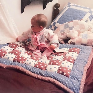 May include: A baby in a pink sweater sits on a blue and white quilted pad with a pink and white patterned border. The pad is on a burgundy bed with a white and blue patterned quilt.