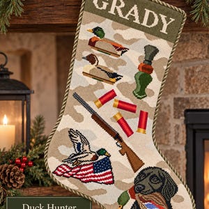 May include: A needlepoint Christmas stocking featuring a camouflage background with the name GRADY at the top. The design includes illustrations of ducks, a duck call, red shotgun shells, a shotgun, an American flag, and a black retriever dog.