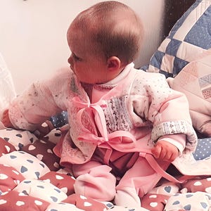 Könnte beinhalten: Ein Baby trägt einen rosa-weißen Pullover mit einem rosa Schleifchen. Das Baby sitzt auf einem herzförmigen, gemusterten Kissen.