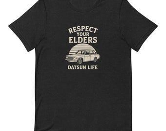 Puede incluir: Camiseta gris oscuro con un diseño gráfico. El diseño presenta una ilustración de coche vintage, un sol y el texto "RESPECT YOUR ELDERS" y "DATSUN LIFE" en un estilo retro. La camiseta está hecha de un material suave.