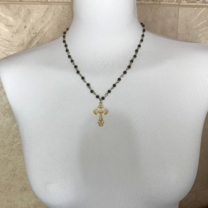 Op de afbeelding: Een gouden kruis hanger ketting met een zwarte kralenketting. Het kruis heeft een gedetailleerd ontwerp en hangt aan een zwarte kralenketting.