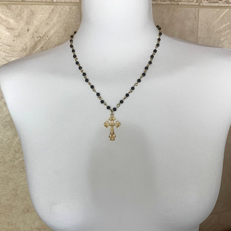 Op de afbeelding: Een gouden kruis hanger ketting met een zwarte kralenketting. Het kruis heeft een gedetailleerd ontwerp en hangt aan een zwarte kralenketting.