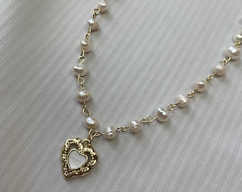 Op de afbeelding: Een delicate gouden ketting met witte parels en een gouden hartvormige hanger. De hartvormige hanger heeft een witte parelmoer inleg.