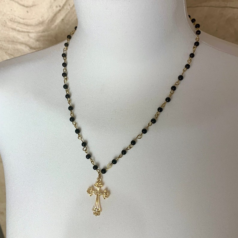 Op de afbeelding: Een gouden kruis hanger ketting met een zwarte kralenketting. Het kruis heeft een gedetailleerd ontwerp en hangt aan een zwarte ketting.