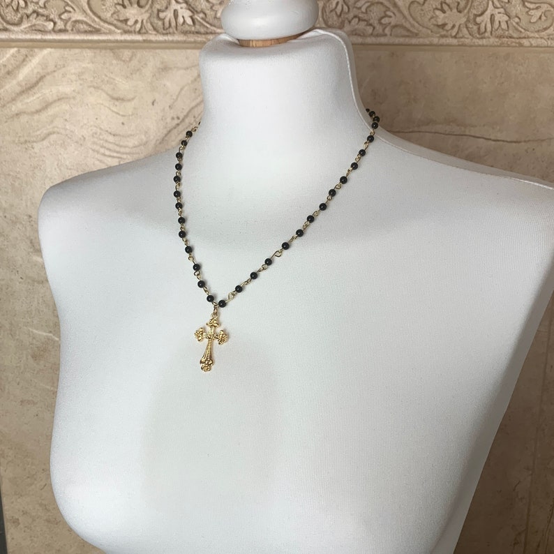 Op de afbeelding: Een gouden kruis ketting met een zwarte kralenketting. De ketting wordt getoond op een wit manikin.