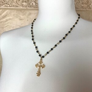 Op de afbeelding: Een zwarte kralenketting met een gouden kruis hanger. Het kruis heeft een gedetailleerd ontwerp.