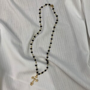 Op de afbeelding: Een gouden kruis hanger ketting met zwarte kralen.