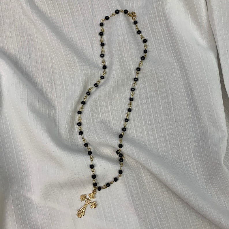 Op de afbeelding: Een gouden kruis hanger ketting met zwarte kralen.