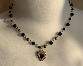Op de afbeelding: Een zwarte kralenketting met een gouden hart hanger. De hart hanger heeft een zwart geëmailleerd midden.
